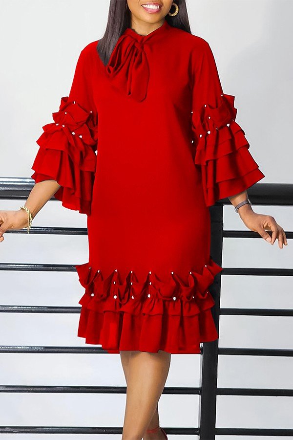 A woman wearing a bright red midi dress with tiered ruffle sleeves, a high neck with a bow, and a matching ruffled hem, standing in front of a white wall with black horizontal bars.