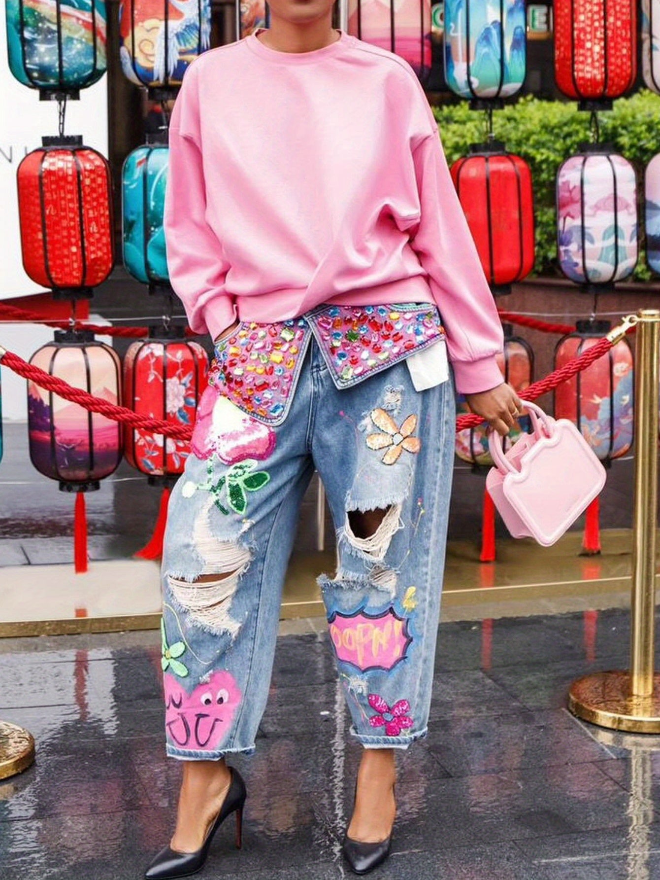 Person wearing a pink sweatshirt and colorful patchwork jeans with a pink handbag, standing in front of decorative lanterns.