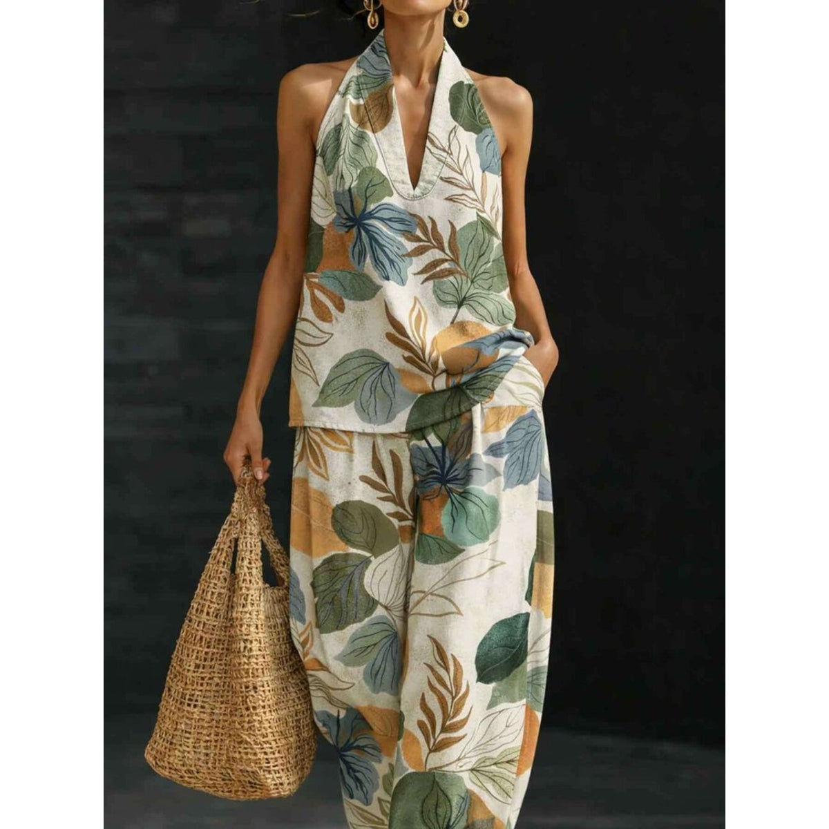 A woman models a sleeveless halter-neck two-piece set with a tropical leaf print in green, blue, and mustard on an off-white background, holding a woven straw bag against a dark studio backdrop.