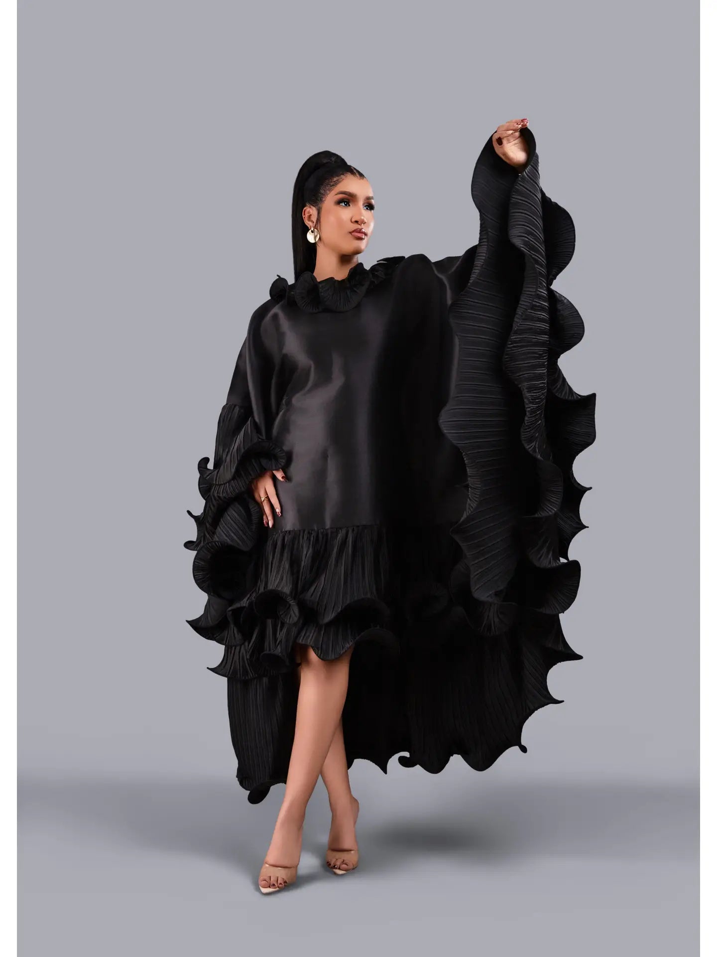 A model wearing a dramatic black ruffled dress with voluminous draped sleeves and a high-low tiered hem, posed against a solid gray studio background.