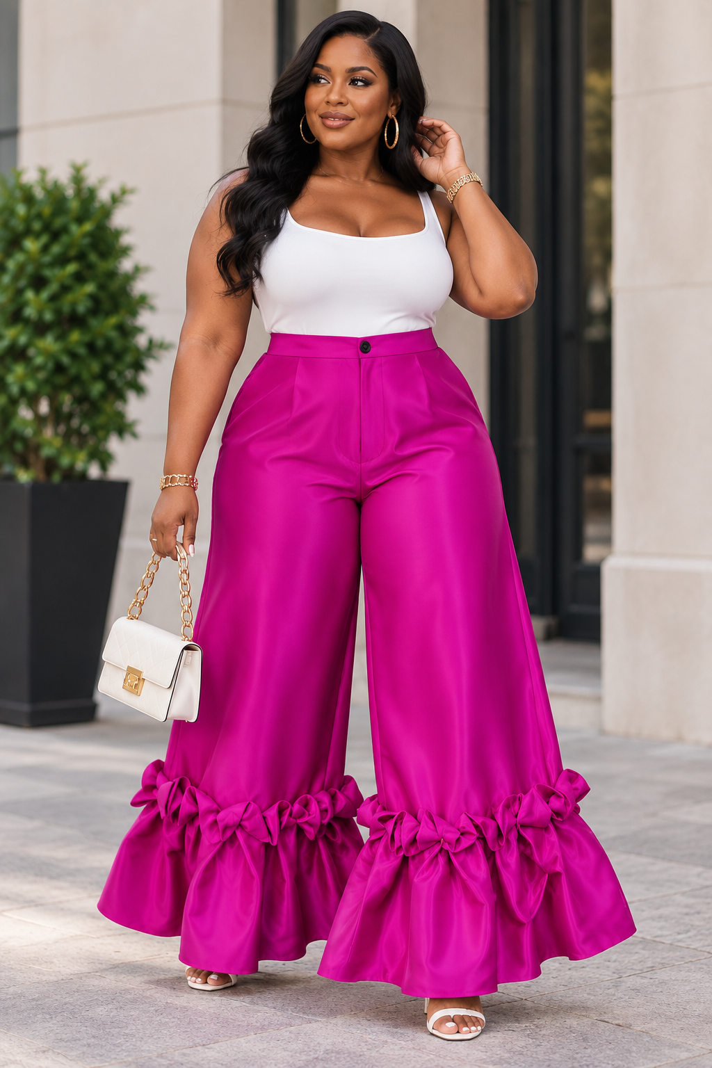 A woman wearing bright fuchsia wide-leg ruffled pants and a white sleeveless top, standing outdoors in front of a building entrance and holding a small white handbag.