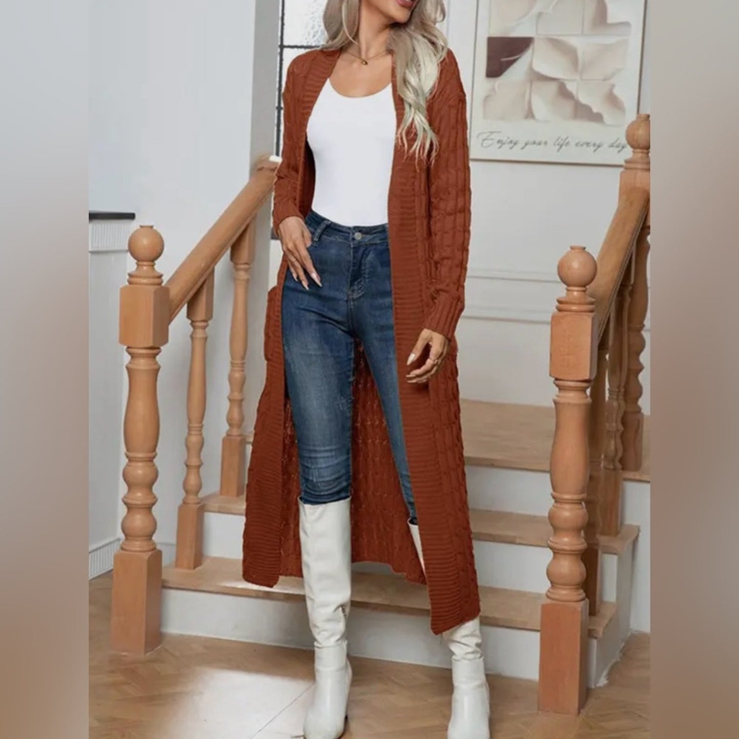 Woman wearing a long rust-colored cardigan, white top, blue jeans, and white knee-high boots standing on a staircase.