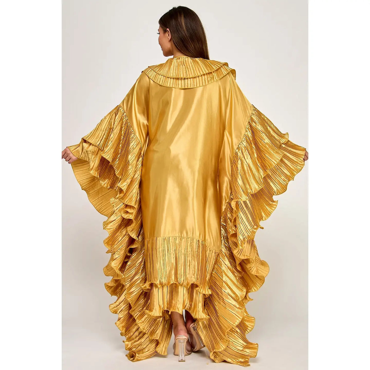 A woman viewed from the back wearing a metallic gold ruffled caftan or poncho-style dress with wide flared sleeves and layered ruffles along the hem, against a plain light gray studio background.