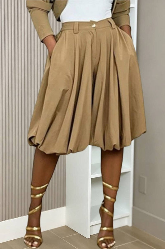 A model wearing a high-waisted, pleated tan midi skirt with a matching belt, styled with a white top, beige cardigan, and gold strappy heels.
