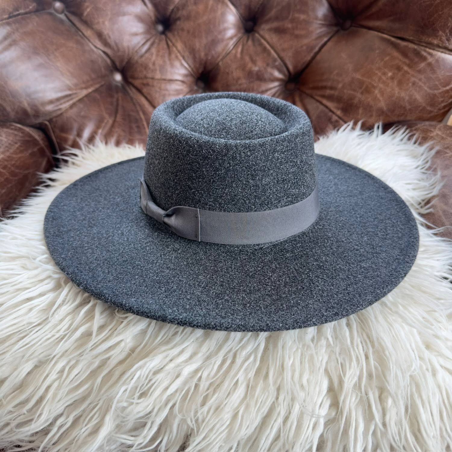 Dark gray fedora hat with a wide brim on a fluffy white surface with a brown leather couch in the background.