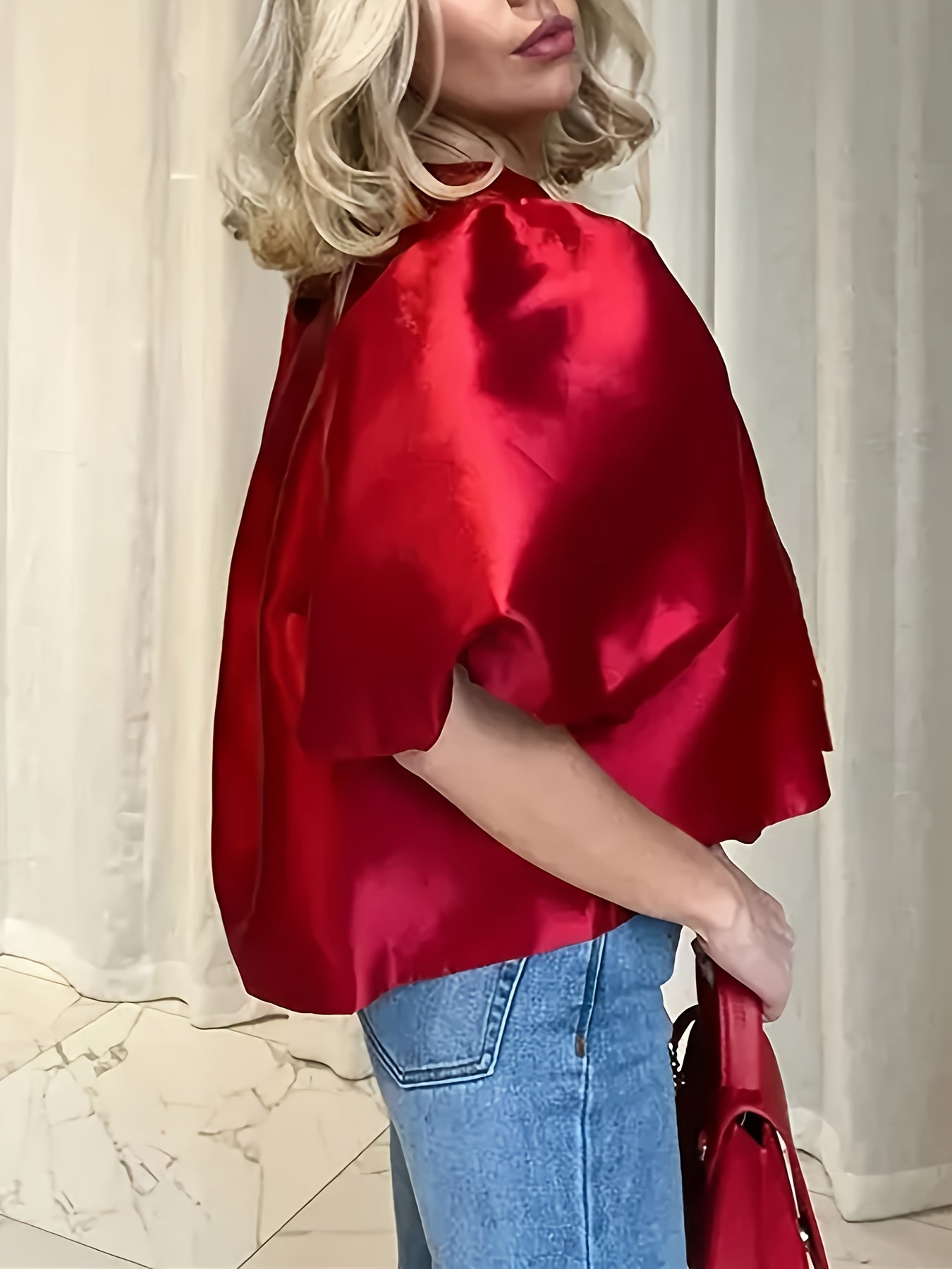 Person wearing a red satin top and blue jeans, holding a red handbag against a white curtain background.