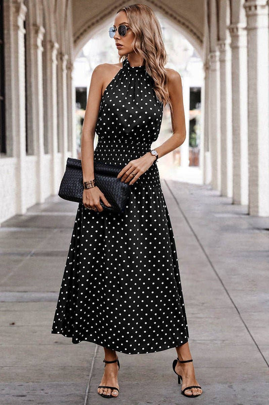 Woman wearing a black polka dot dress with sunglasses and clutch in an outdoor setting.
