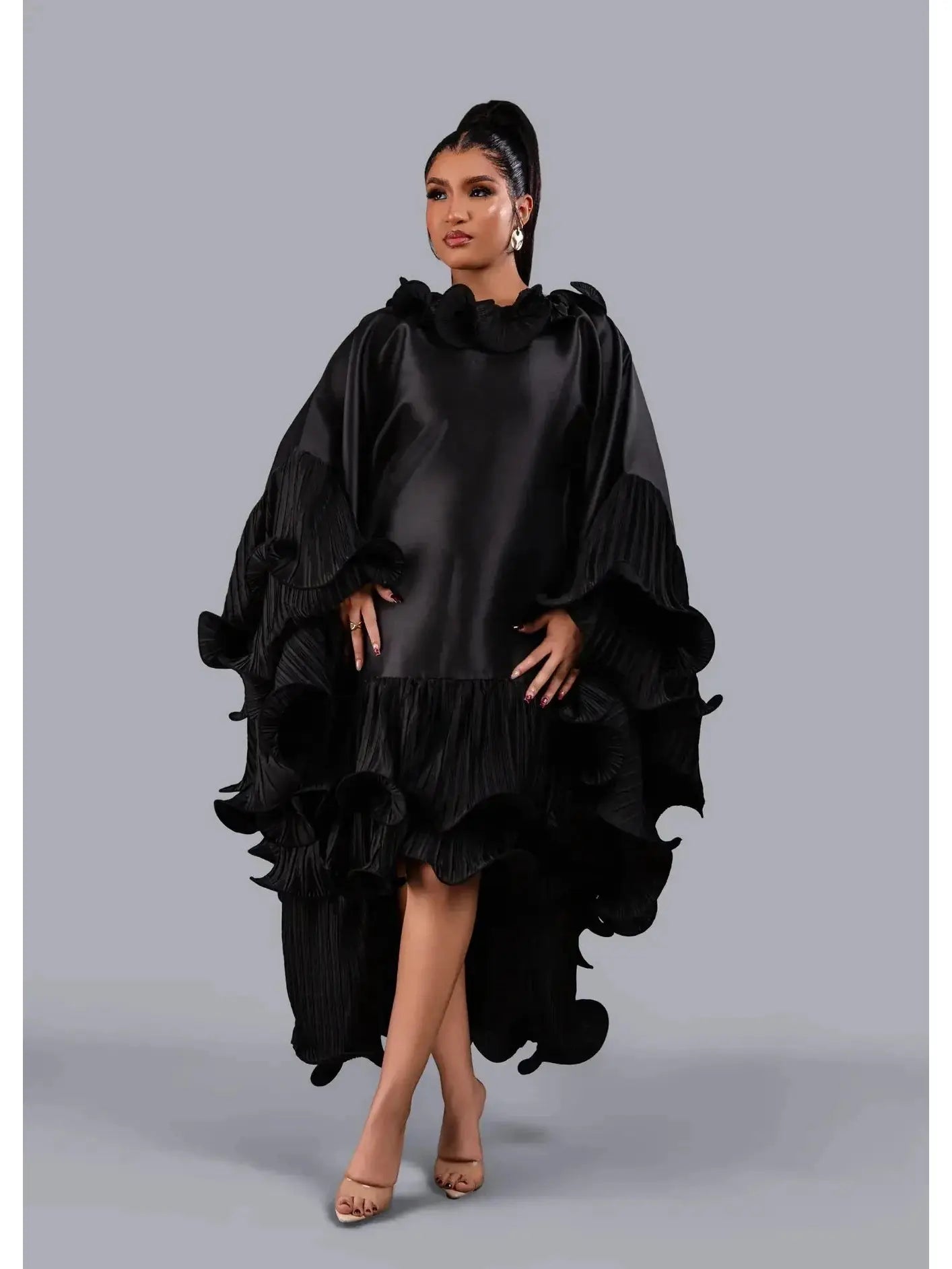 A woman modeling a black high-low dress with dramatic layered ruffles and a ruffled collar, photographed against a solid gray studio background.