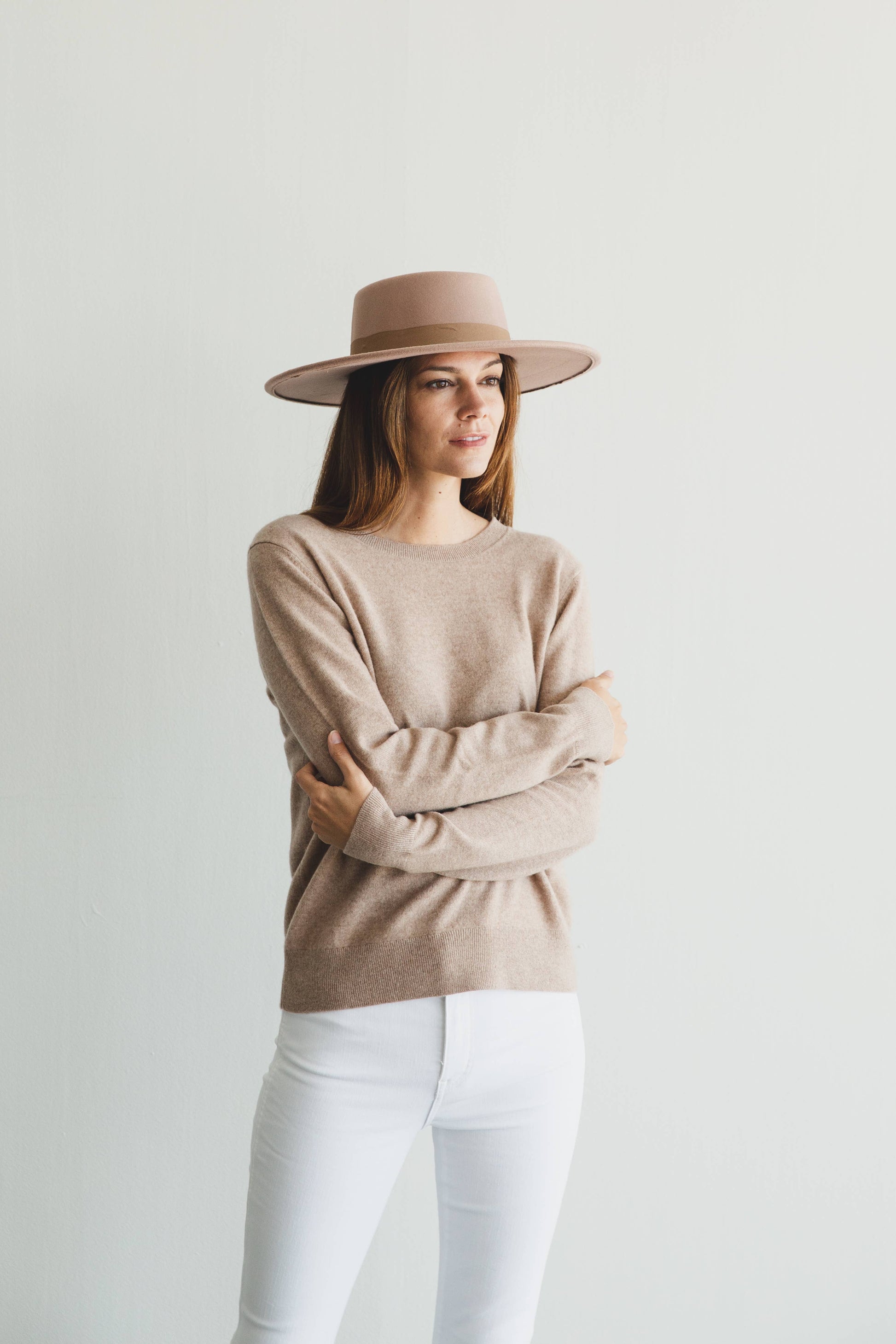 Woman wearing a beige sweater and hat with white pants against a light background