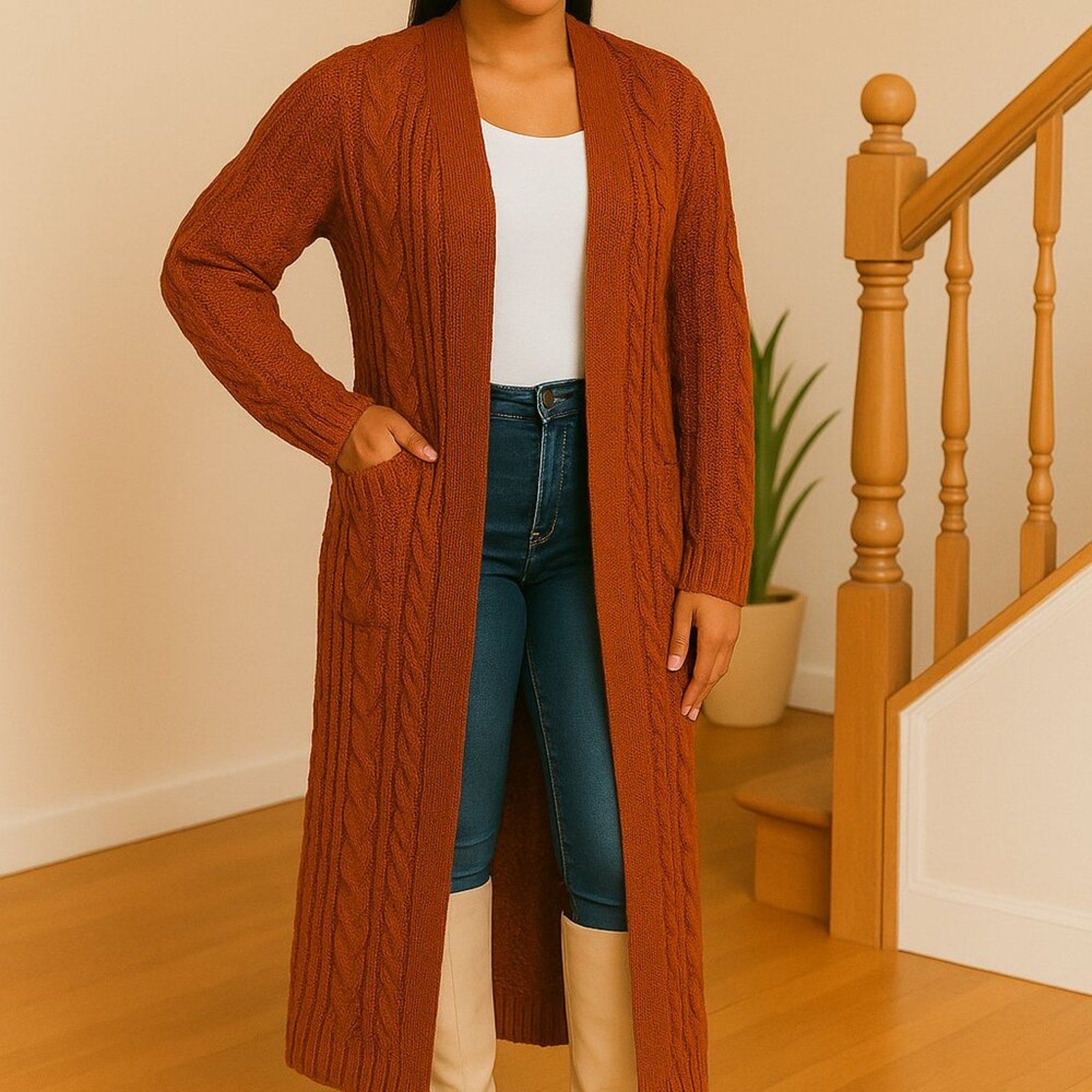 Person wearing a long rust-colored cardigan over a white top and blue jeans, standing in a home setting with wooden stairs and a plant.