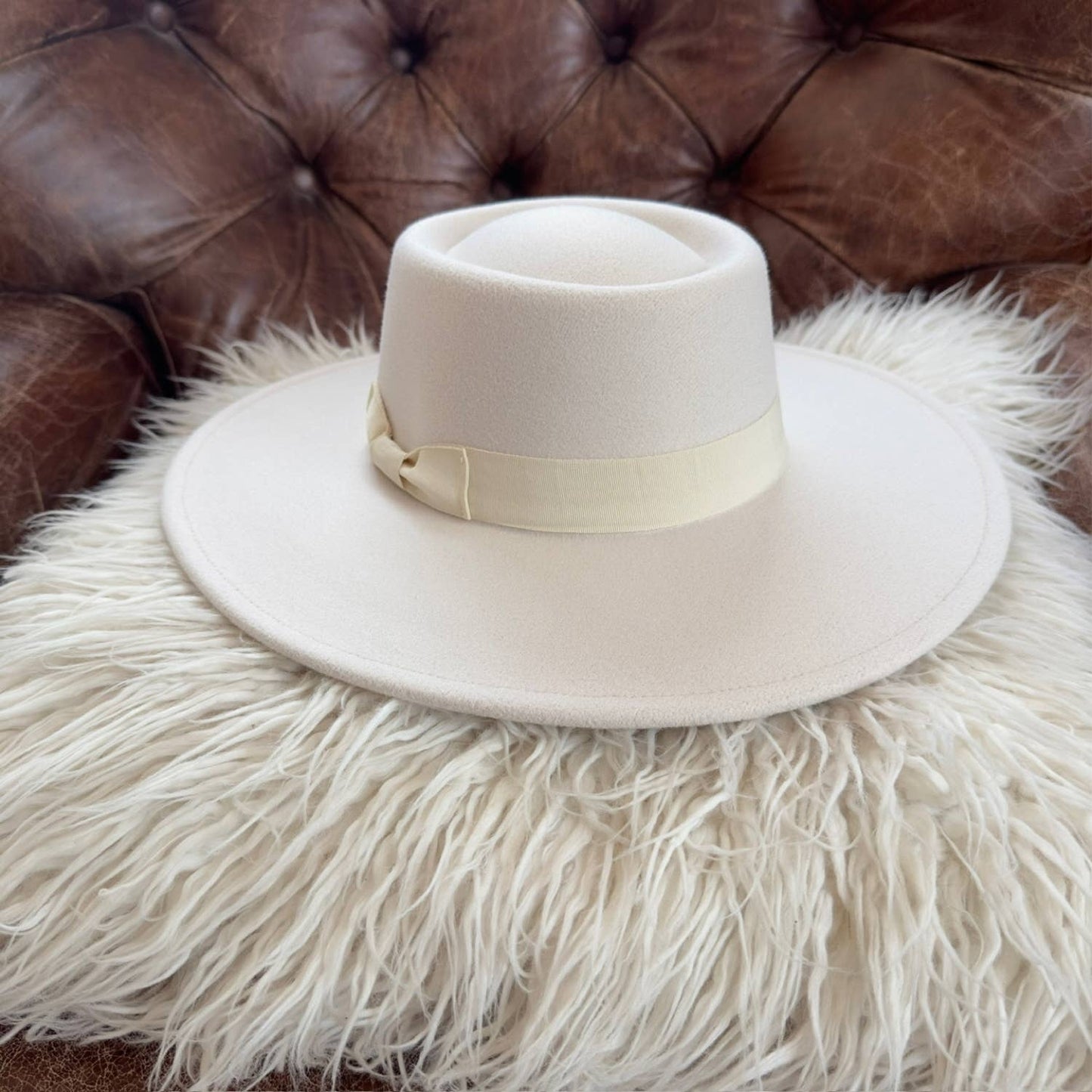 White fedora hat with a ribbon on a fluffy white surface with a brown leather couch in the background.
