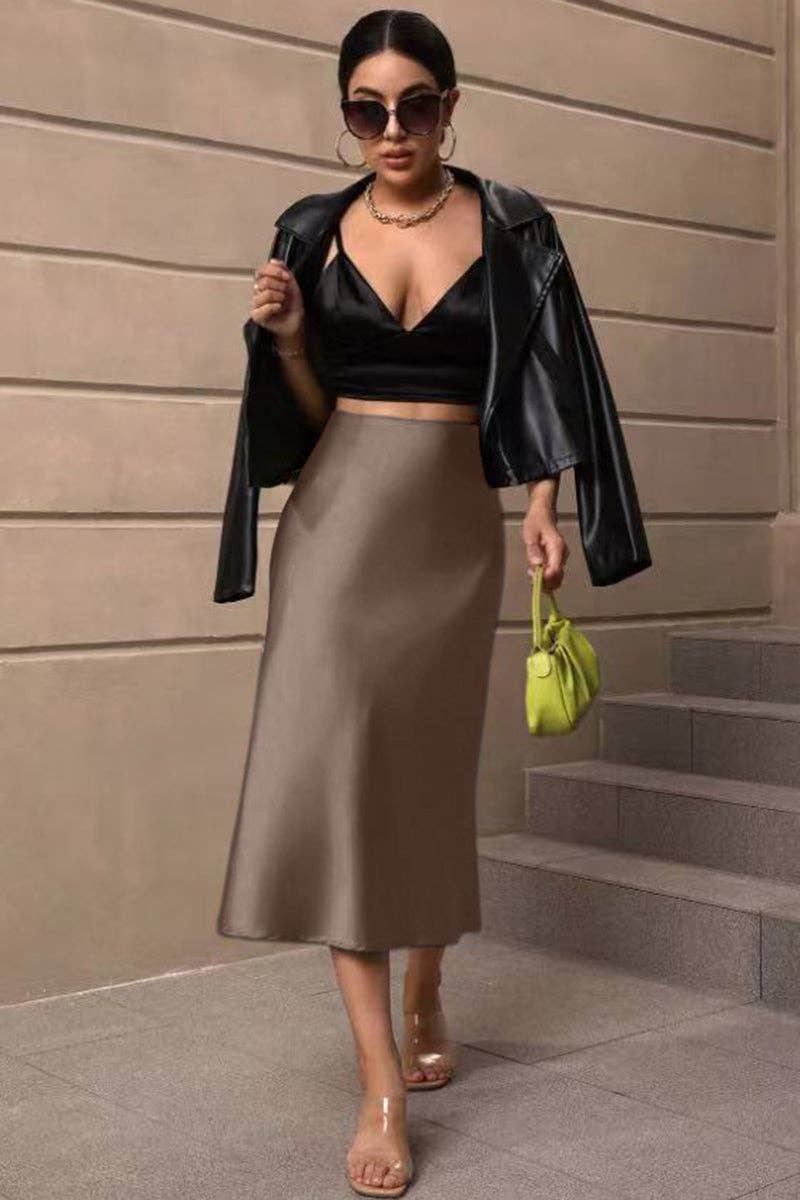 Woman in black leather jacket, black top, and brown satin skirt holding a green handbag against a beige wall.