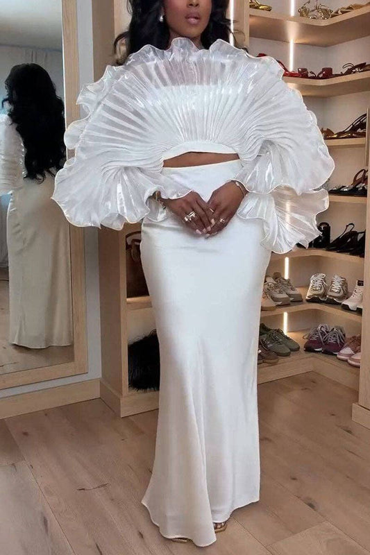 A woman wearing a white two-piece dress with a dramatic ruffled overskirt and a fitted satin skirt, standing in a boutique with wooden floors and shoe shelves in the background.