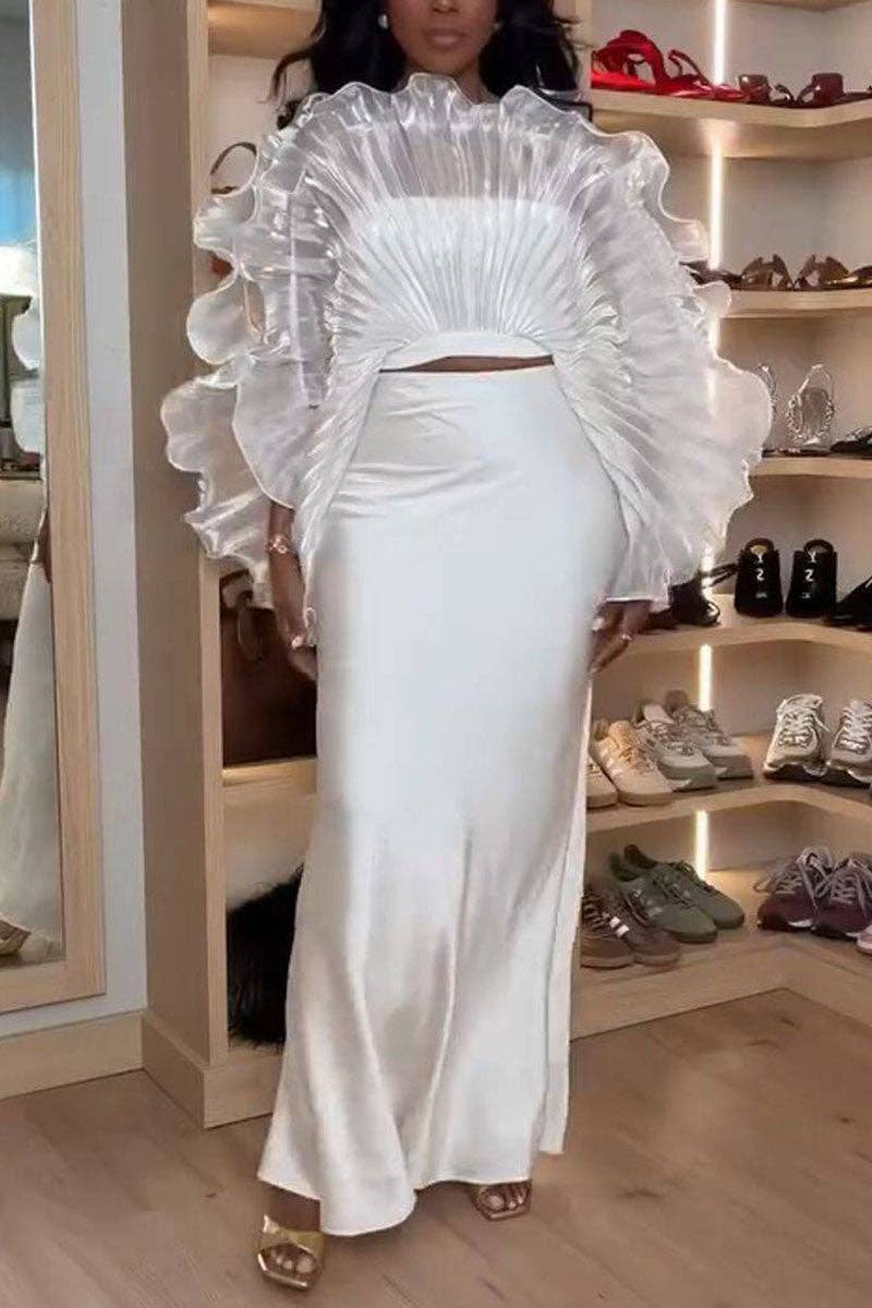 A woman models a white two-piece satin outfit featuring a high-neck crop top with dramatic layered ruffles and a matching fitted maxi skirt, set against a boutique background with shoe shelves.