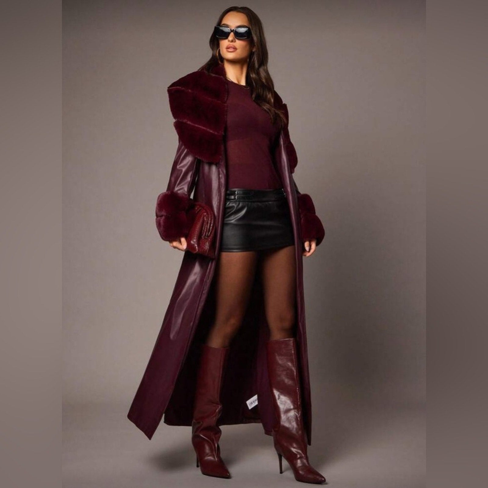 Woman wearing a long burgundy coat, top, and boots against a gray background