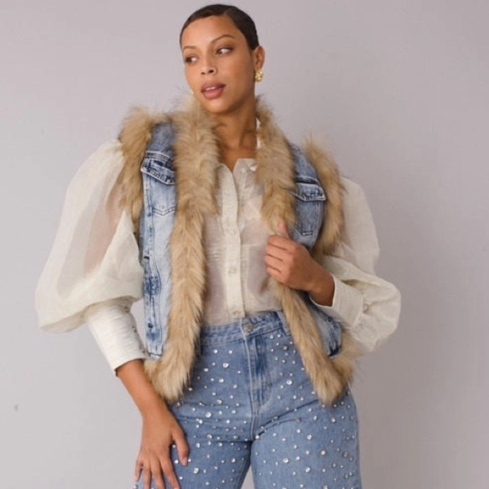 Woman wearing a denim vest with fur trim and polka dot jeans on a plain background