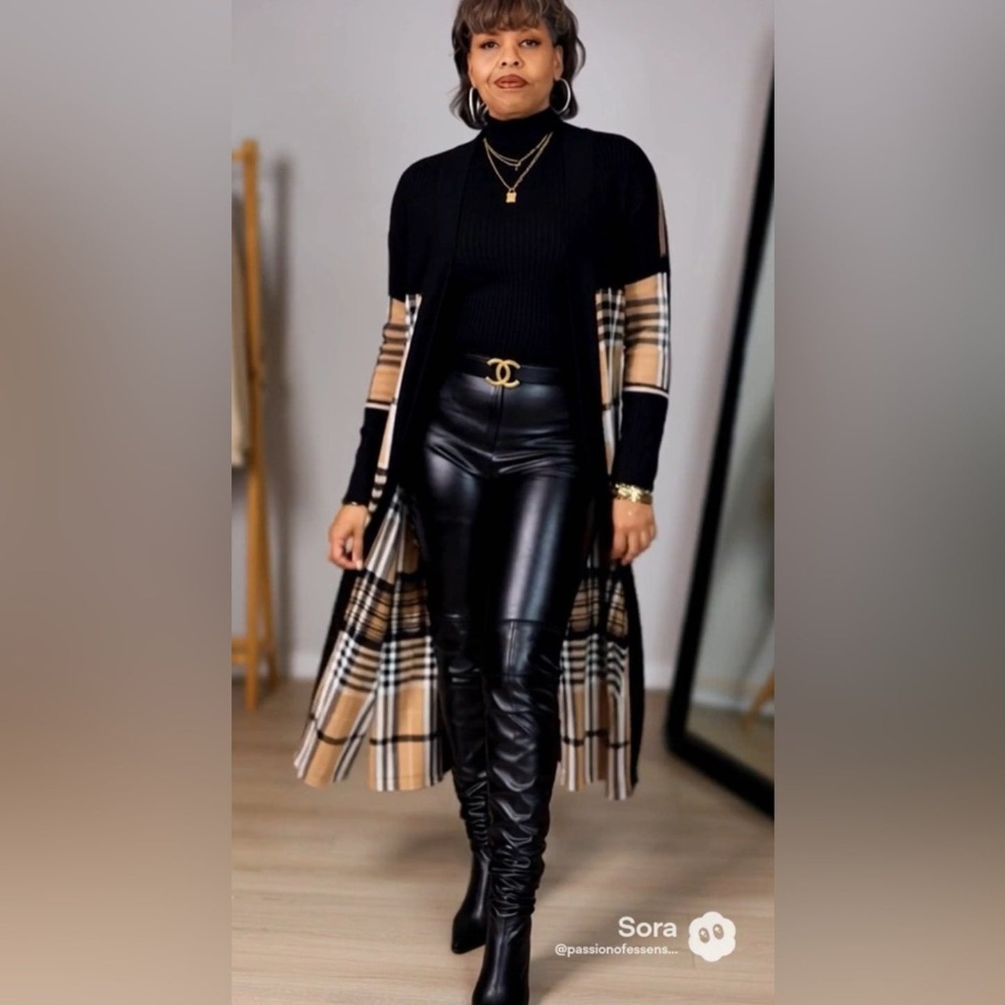 Woman wearing a black outfit with plaid sleeves and a long coat, standing indoors.