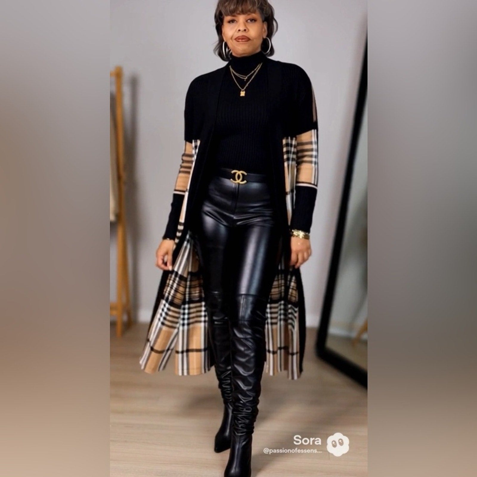 Woman wearing a black outfit with plaid sleeves and a long coat, standing indoors.