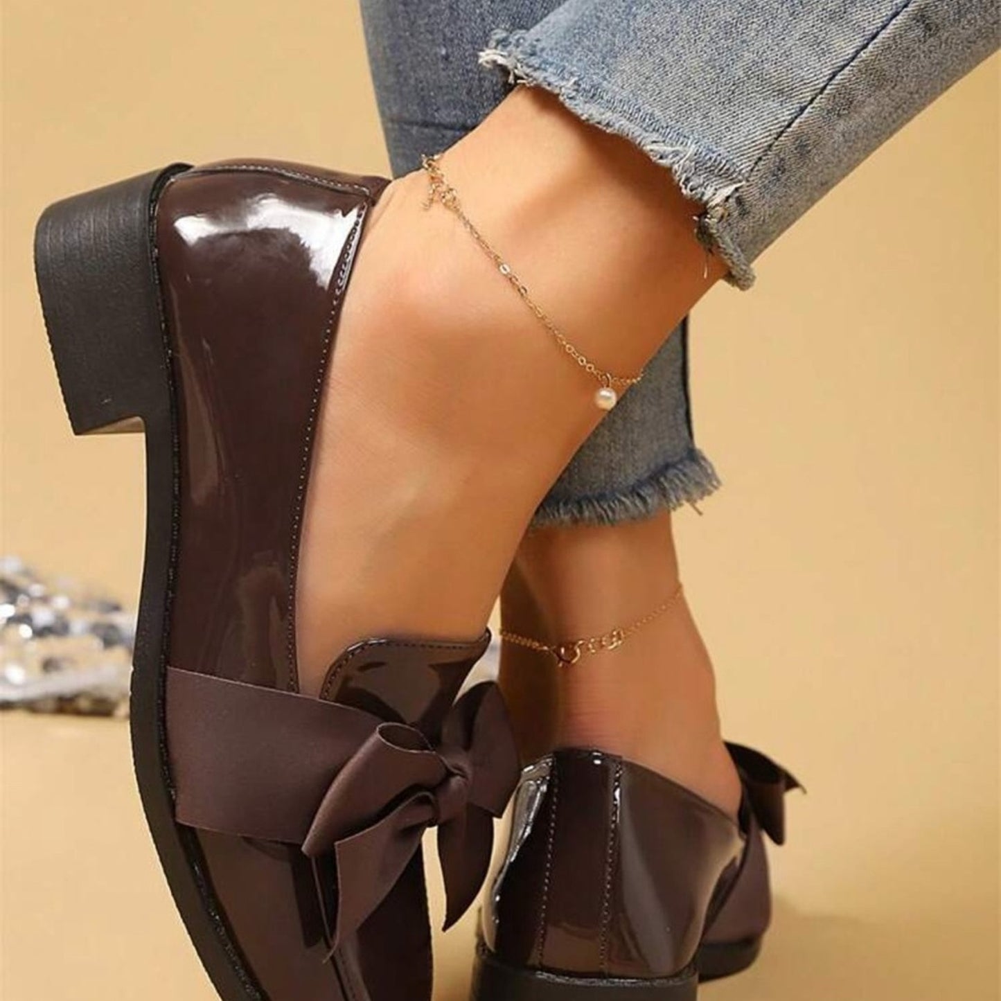 Brown patent leather shoes with a bow detail worn by a person in denim jeans.