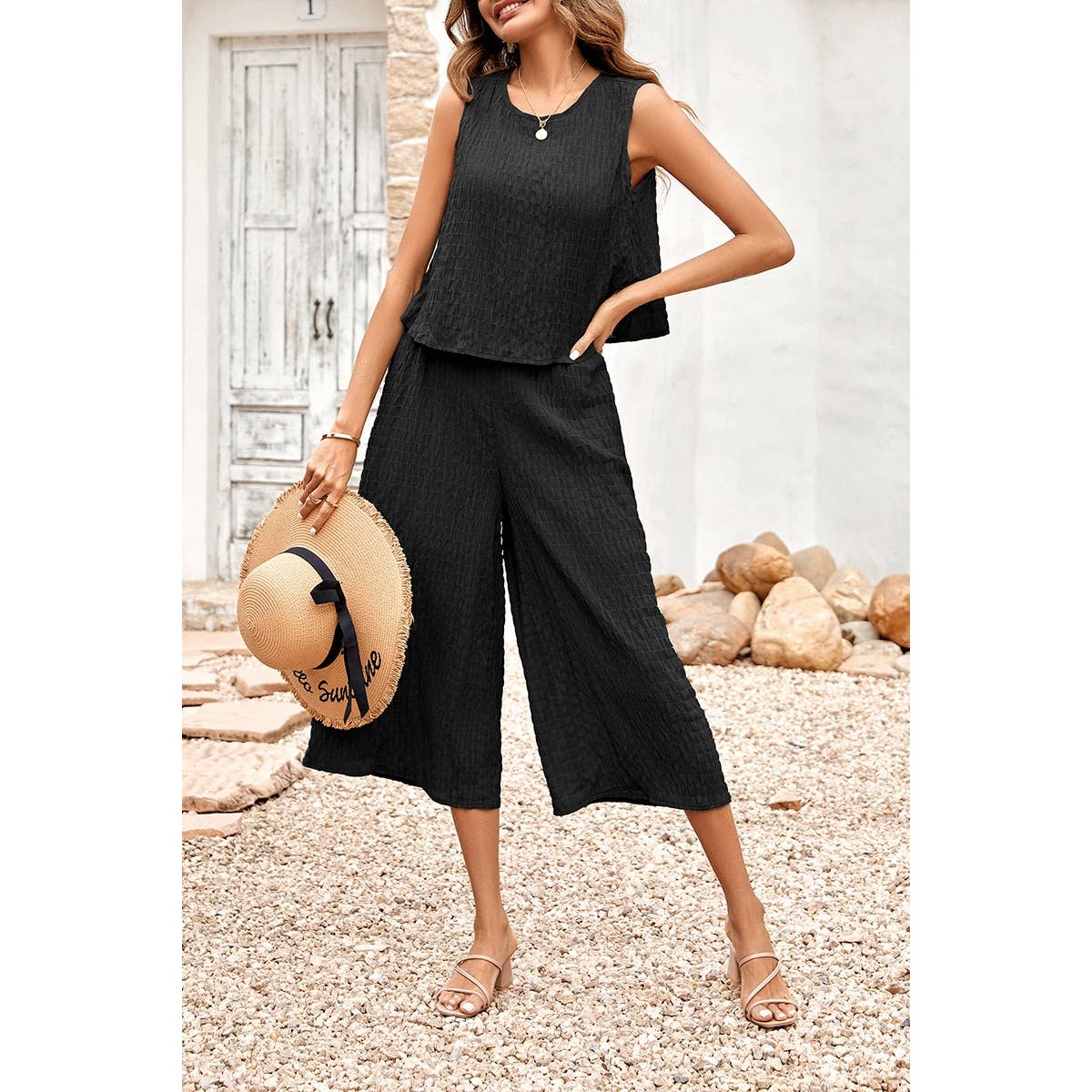 Woman in black sleeveless top and wide-leg pants holding a straw hat outdoors.