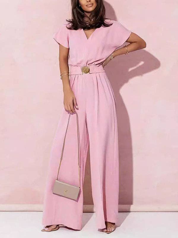 Woman wearing a pink jumpsuit with a belt and holding a silver handbag against a pink background