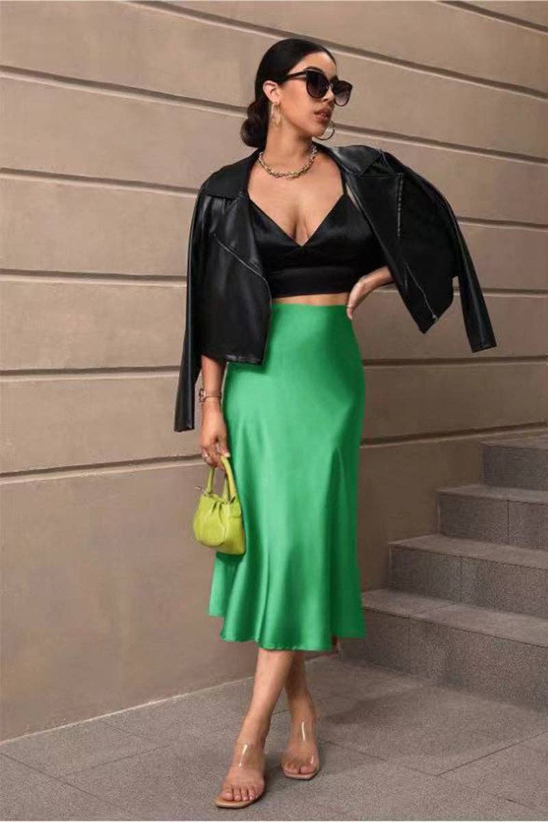 Woman in black jacket, black top, and green skirt standing against a beige wall.