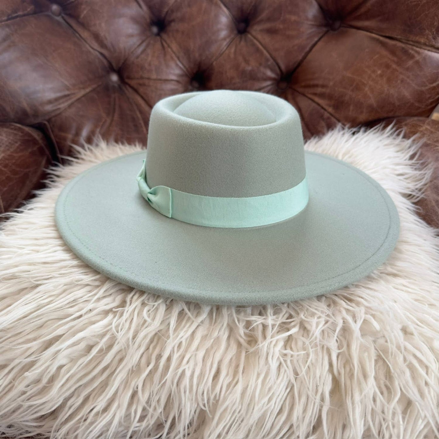 Light blue fedora hat with a green band on a fluffy white surface with a brown leather couch in the background.