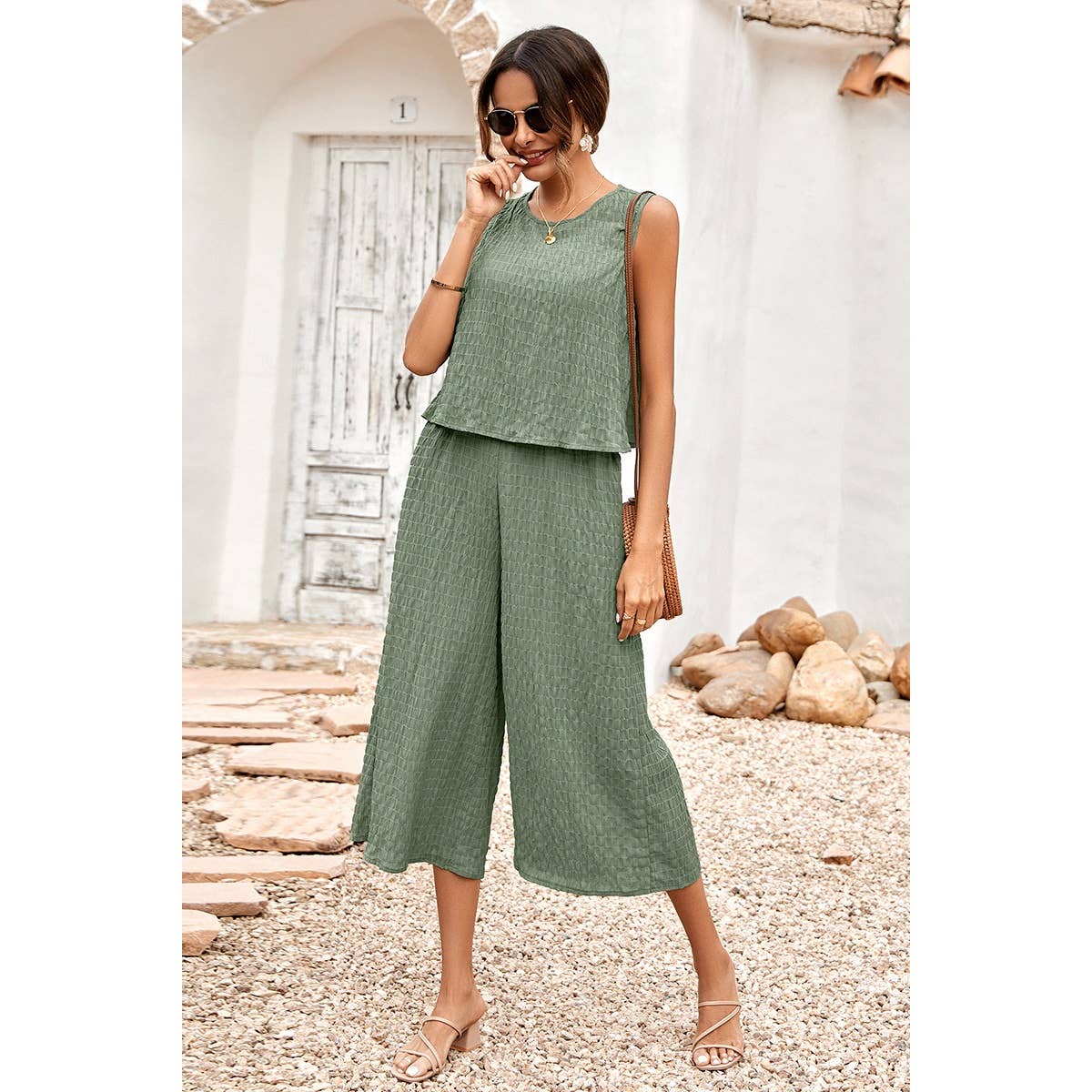 Woman wearing a green sleeveless top and wide-leg pants standing in front of a white wall with wooden elements.