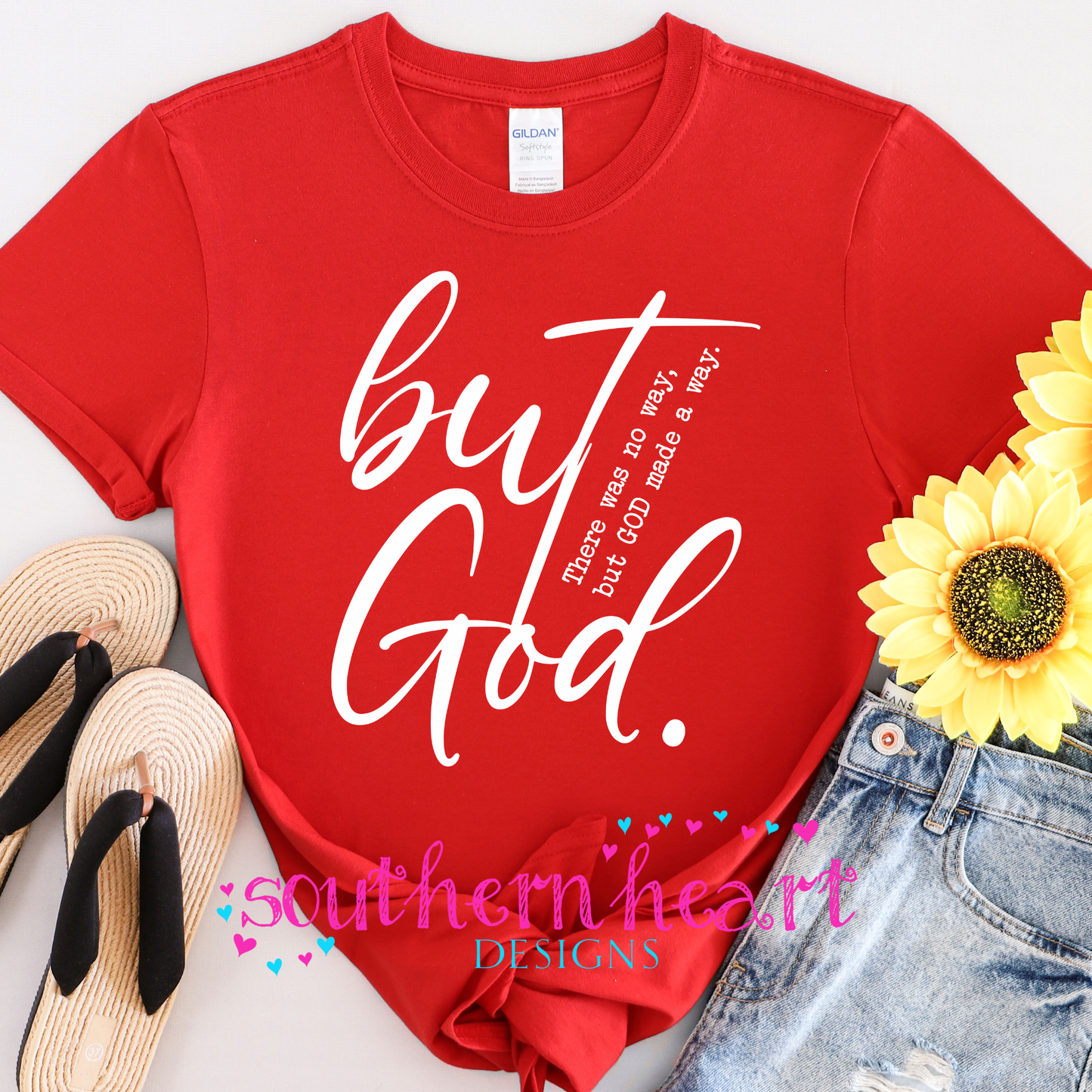 Red t-shirt with white text 'but God' and 'southern heart DESIGNS' on a white background with sunflower and denim jeans.