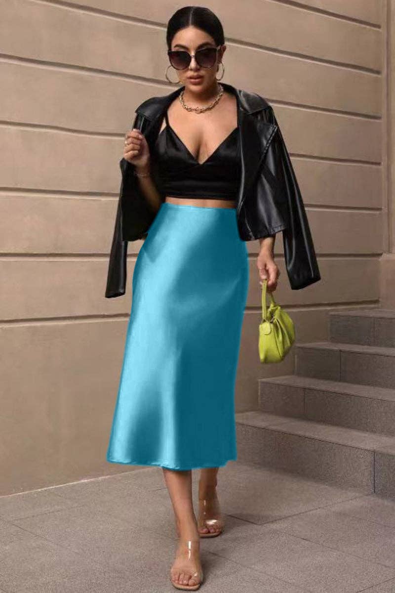 Woman in black leather jacket, black top, and blue satin skirt holding a green handbag against a beige wall.