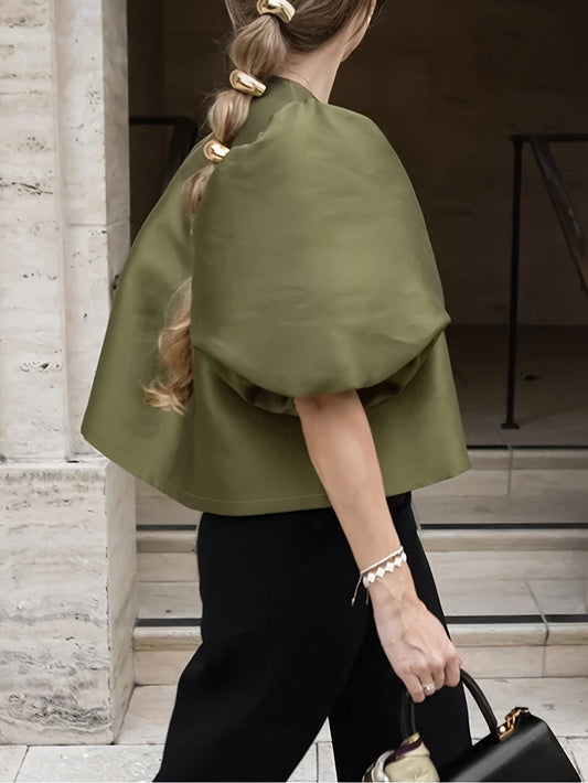 Person wearing a green cape and black pants, holding a black handbag, standing in front of a stone building.