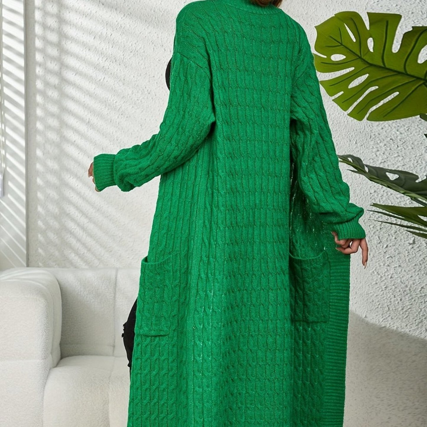 Person wearing a long green knitted cardigan in a room with a white sofa and plant.