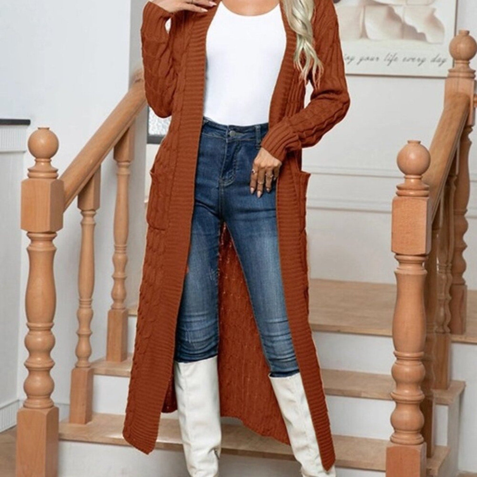 Person wearing a long brown cardigan, white top, blue jeans, and white boots on wooden stairs.