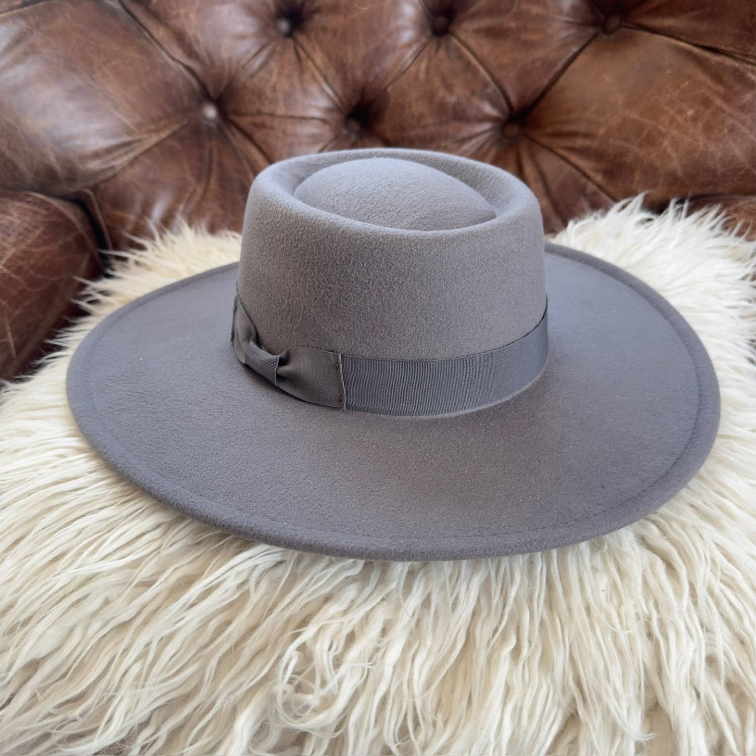 Gray fedora hat with a bow on a fluffy white surface with a brown leather couch in the background