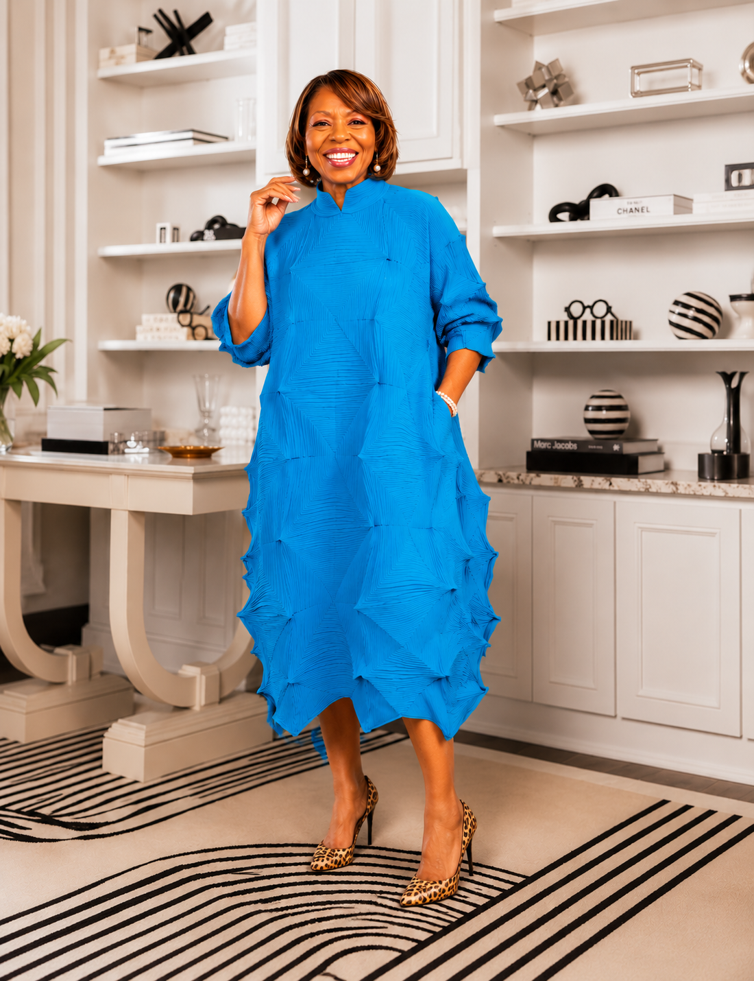 Woman in a blue dress standing in a stylish room with shelves and decor.