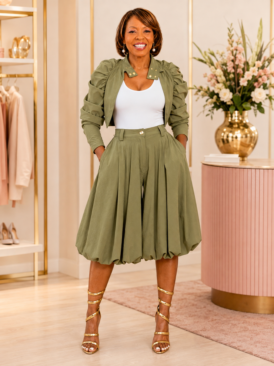 A woman models an olive green two-piece set consisting of a cropped jacket with puffed sleeves and matching high-waisted pleated skirt, styled over a white tank top and gold lace-up heels in a boutique setting.