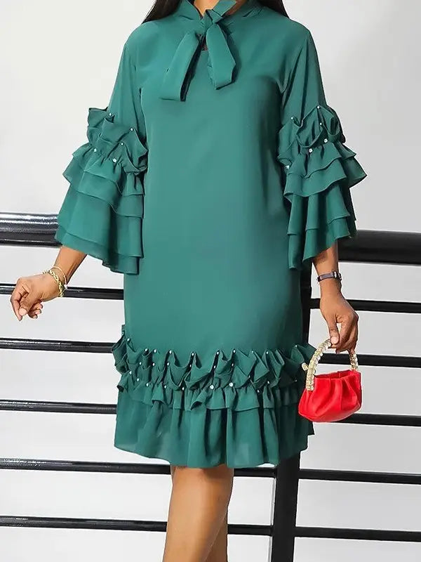 A woman wearing a teal green tiered ruffle dress with a bow tie neckline, holding a small red handbag in front of a black metal railing.