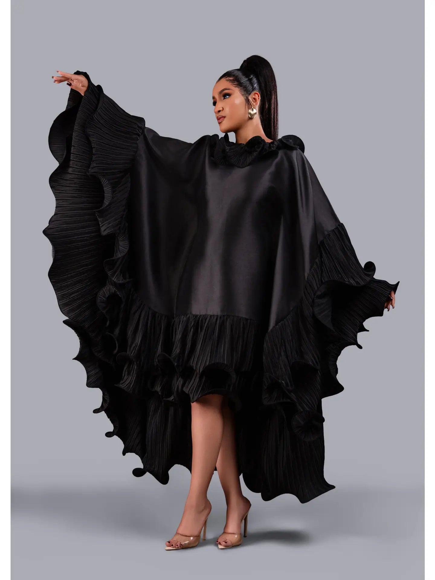 A woman modeling a dramatic black high-low dress with wide, pleated ruffled sleeves and a ruffled hemline, posed against a solid gray studio background.