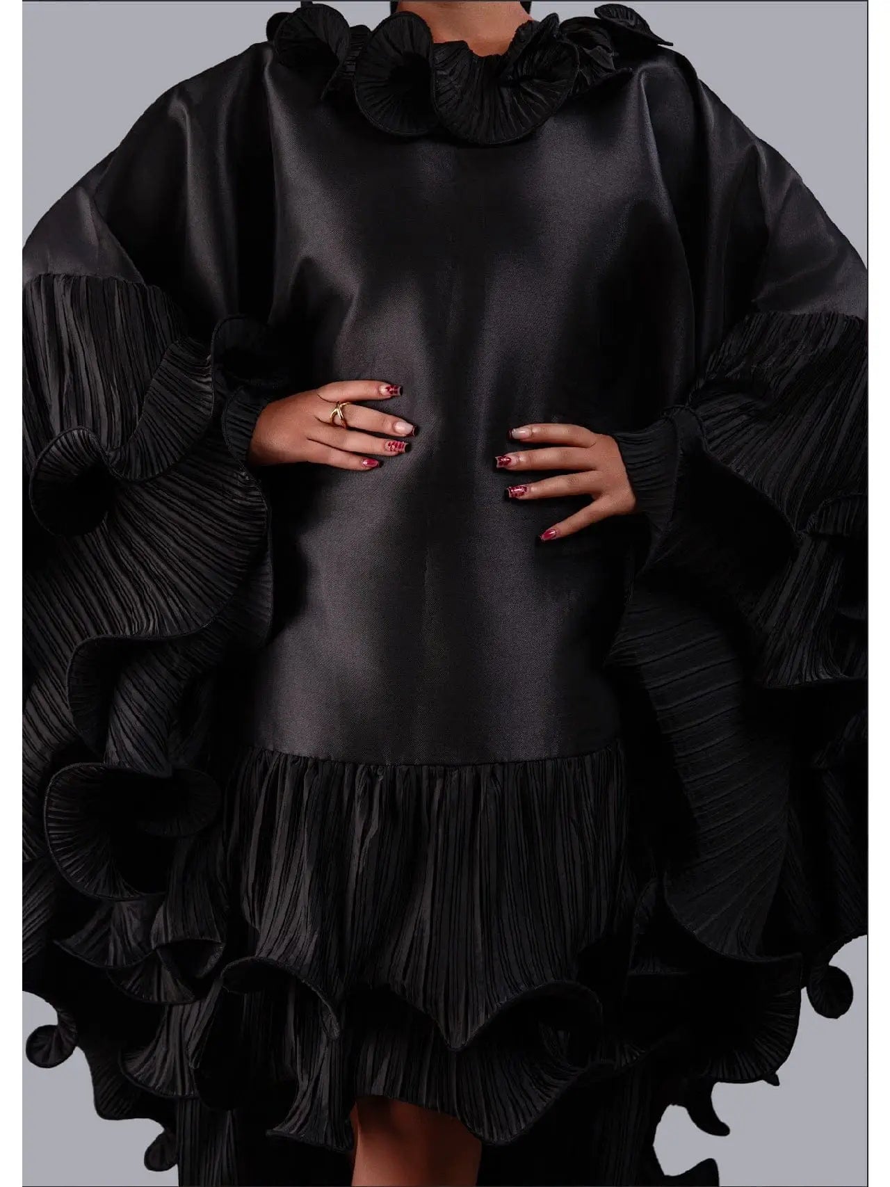 A black satin-like mini dress with dramatic pleated ruffle details, including a high ruffled collar, voluminous sleeves, and a tiered ruffle hem, shown on a model against a solid gray background.