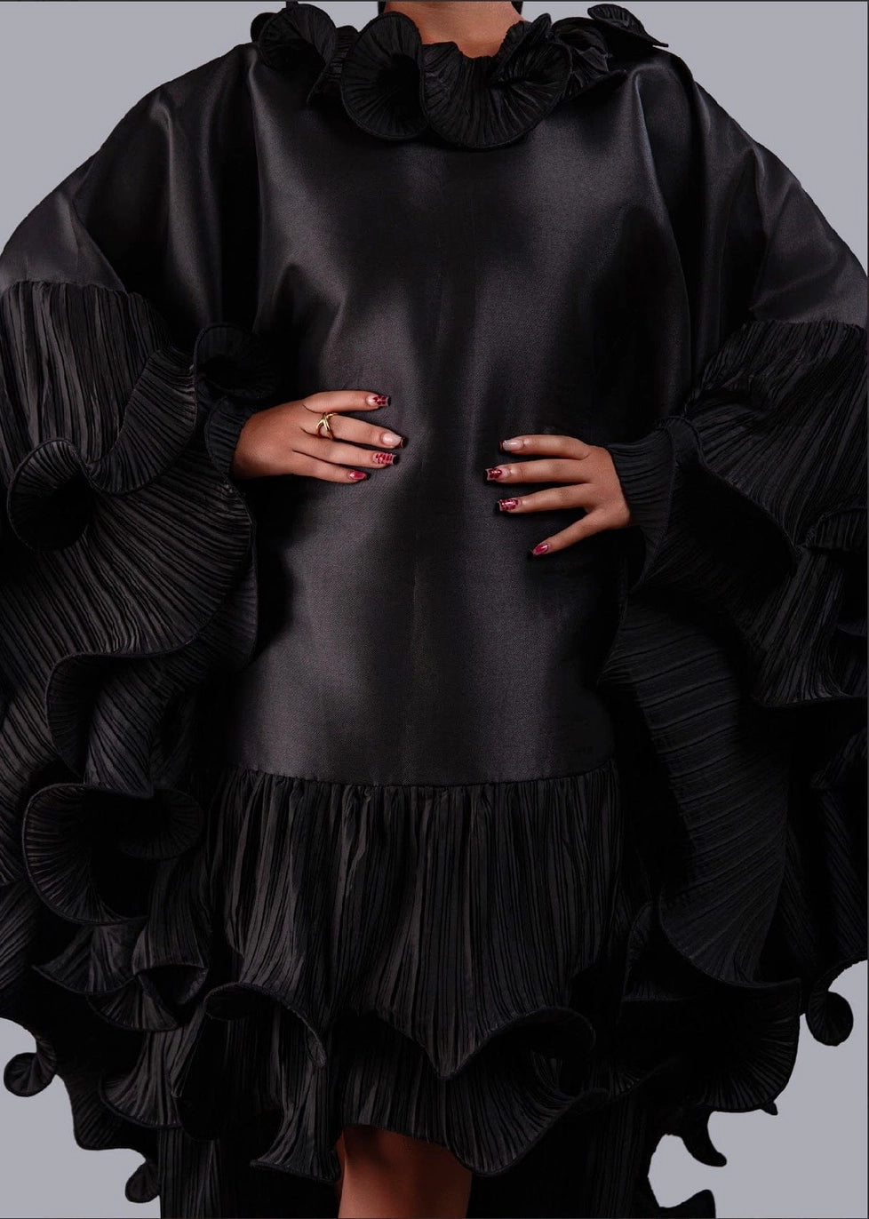 A close-up of a model wearing a black ruffled tunic dress with pleated wide sleeves and a ruffled high neckline, set against a solid gray background.