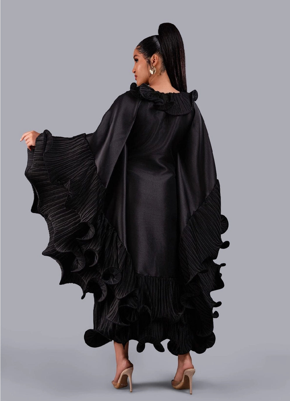 A woman modeling a black flamenca dress with dramatic pleated ruffle details, shown from the back against a solid gray studio background.