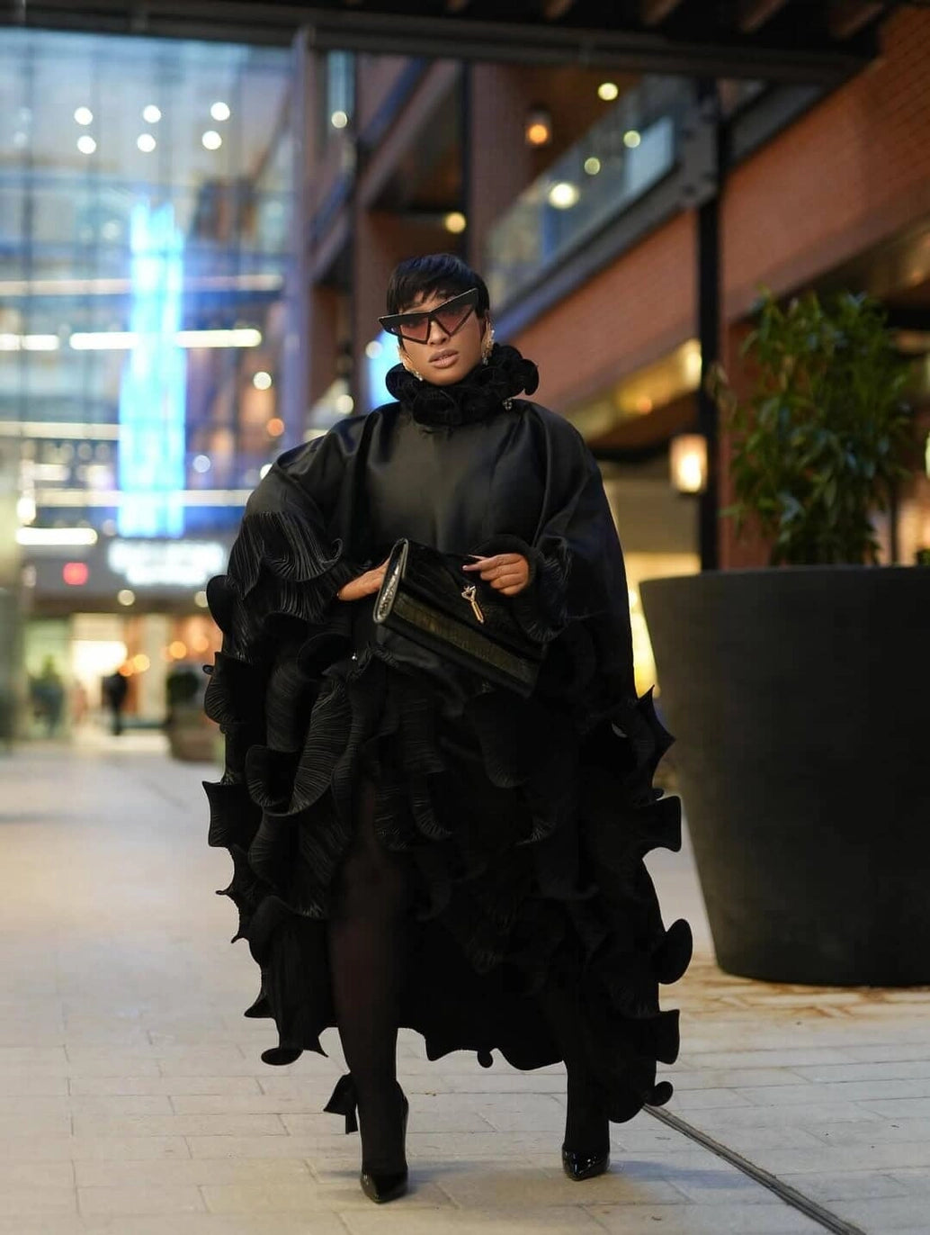 A person walking outdoors at night wearing an elaborate black ruffled cape-style dress or outfit with a high ruffled collar, black tights, black pointed shoes, and carrying a black handbag.