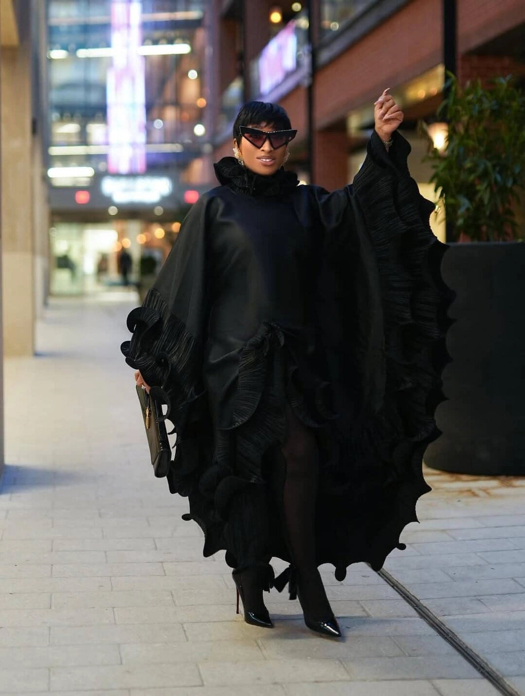 A model walking outdoors in a black ruffled cape-style dress with dramatic layered edges, black tights, black pointed heels, and dark sunglasses on a city sidewalk at night.