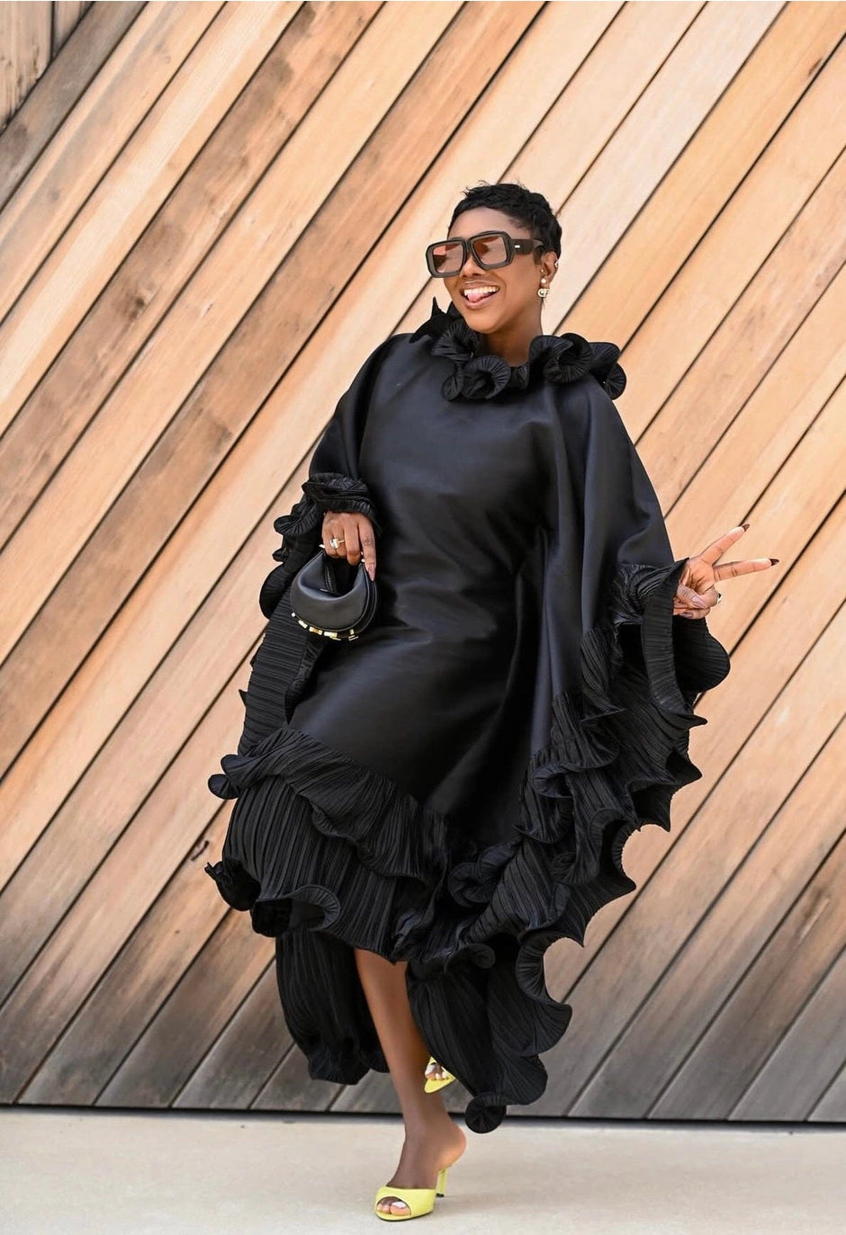 A woman wearing a dramatic black ruffled dress with wide draped sleeves, holding a small black handbag, posing in front of a diagonal wood-paneled wall.