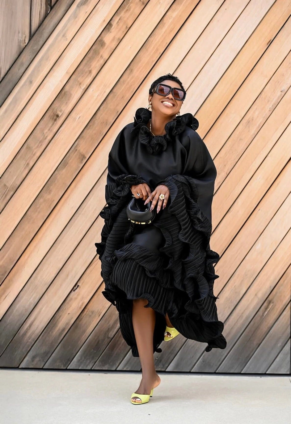 A woman wearing a dramatic black ruffled dress with oversized ruffle details, sunglasses, and neon yellow slide sandals, posing in front of a diagonal wood-paneled wall.