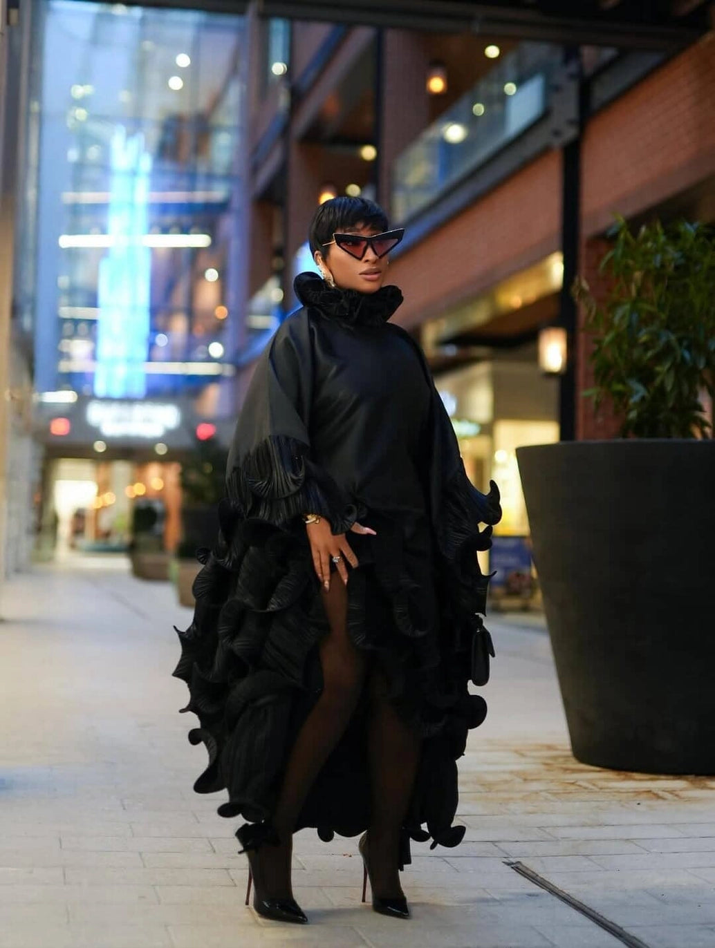 A woman wearing a dramatic black ruffled poncho-style dress with sheer tights and black pointed heels, standing in a modern outdoor shopping plaza at night.