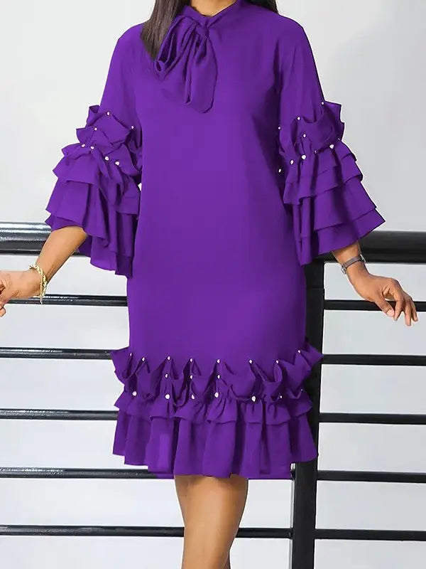 A woman wearing a purple knee-length dress with a high neck, front bow tie, and dramatic tiered ruffle sleeves and hem, posed in front of a white wall and black railing.