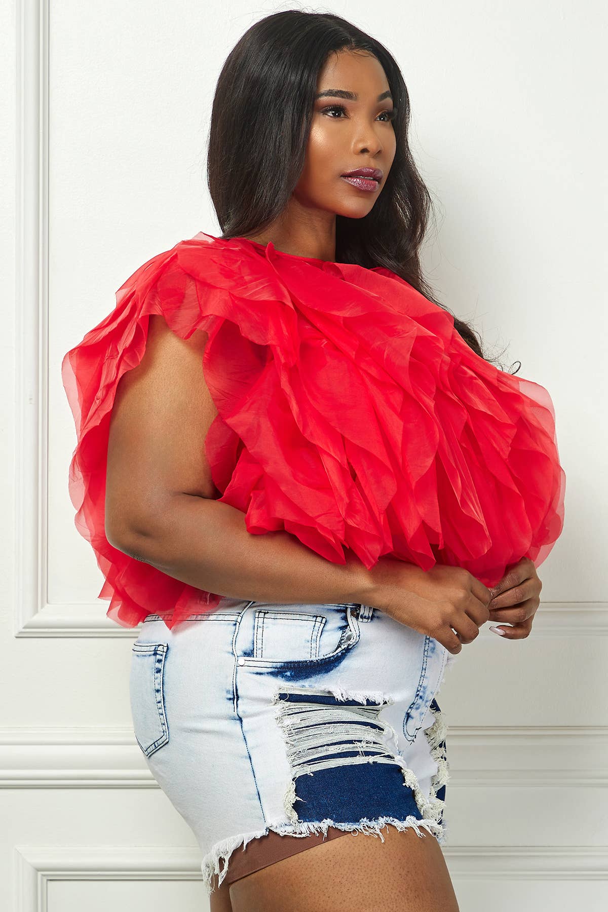 Woman wearing a red ruffled top and denim shorts against a white wall