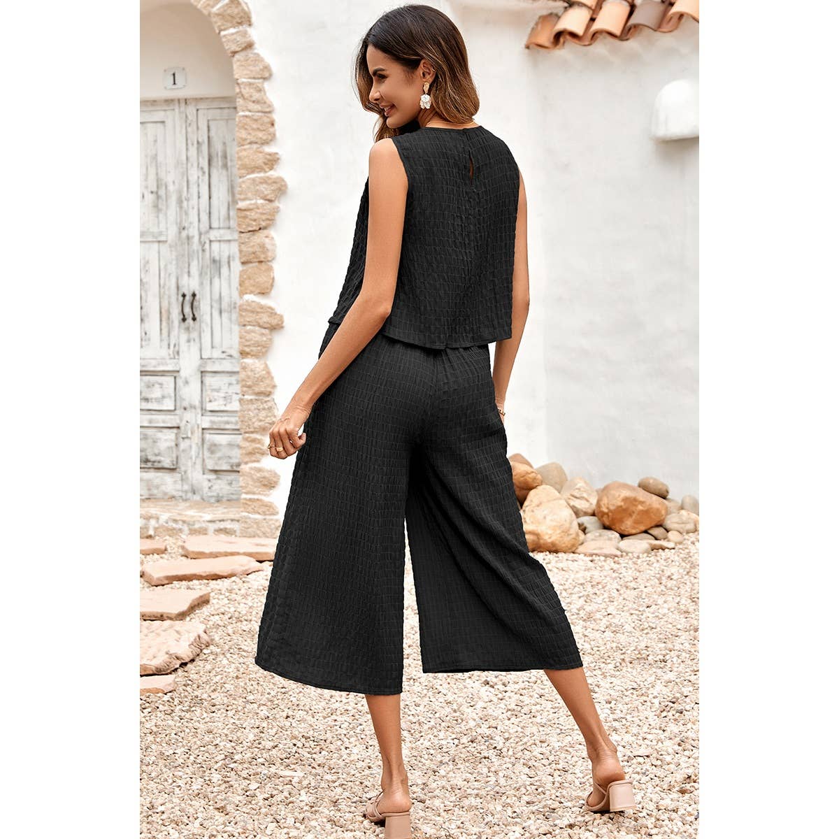 Woman wearing a black sleeveless top and wide-leg pants standing in a rustic outdoor setting.