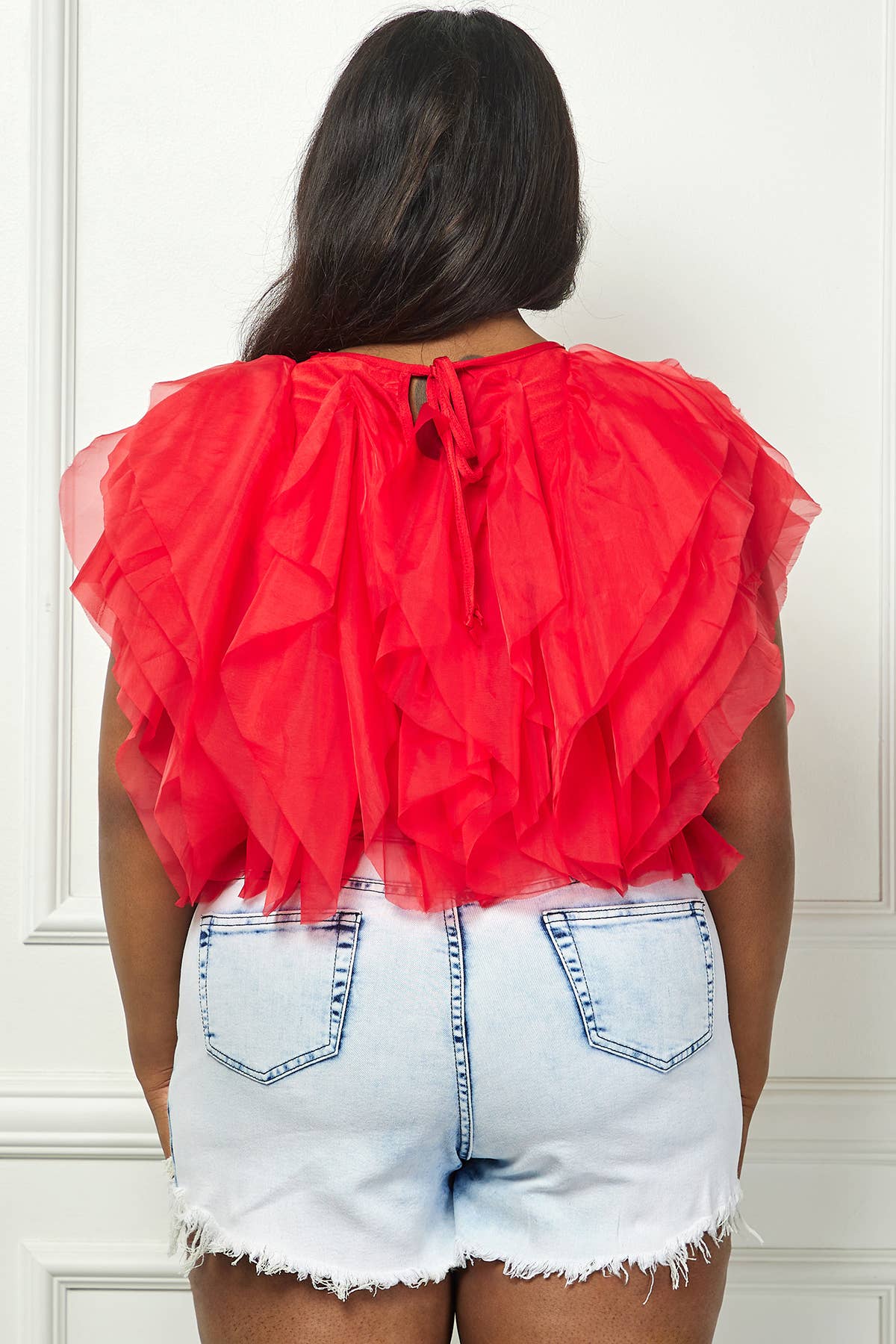 Person wearing a red ruffled top and denim shorts against a white wall.